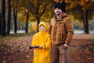 Child wearing a bright yellow jacket holding a flashlight and standing next to a man. Autumn trees and leaves in the background