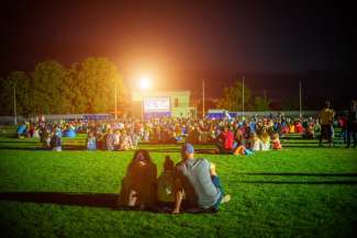 Crowd of people watch movie outside at a baseball field.