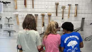 Three members of artist studio summer camp stand facing a wall of jewelry making tools
