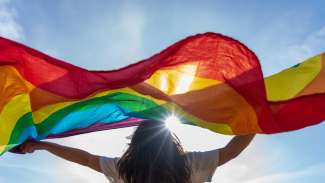person waiving a pride flag with the sun and sky in the background