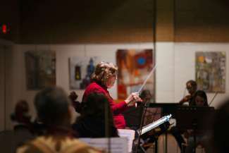 White woman with brown hair conducting a youth orchestra in a gallery, standing in front of a music stand with a baton raised. 