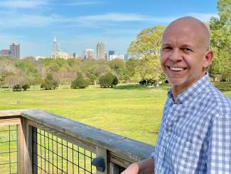 Smiling white man in a blue gingham shirt, leaning on a rain in a park setting with downtown raleigh skyling in the background. 