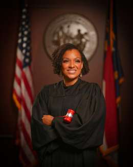 Portrait of Judge Parker in court room with Seal of NC on the wall behind her. An American flag is to her left and a NC flag to her right.