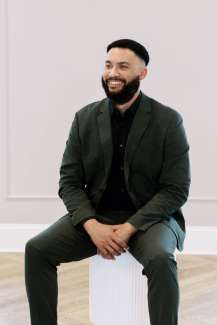 Young Black man dressed professionally in a grey suit and black tie, sitting and smiling broadly. 