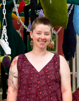 A young white woman with a shaved head and a big smile stands in a studio with colorful artork. 