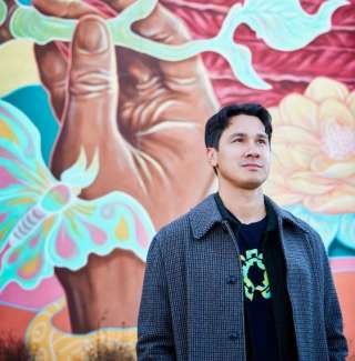 Young man with black hair stands in front of a colorful mural depicting a large brown hand. 