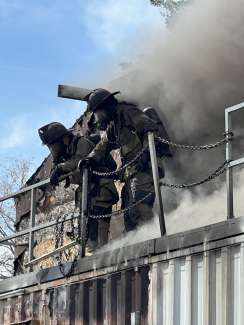 Two firefighters stand outside of a building with black, thick smoke coming out of it.