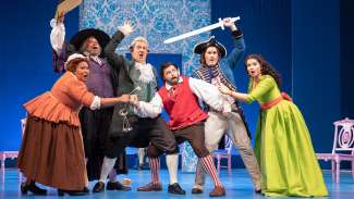 Characters on a stage holding various props huddled together looking in the same direction with surpised looks on faces