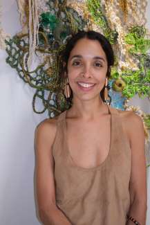 A woman with dark hair and gold earrings poses against a wall with fiber art.