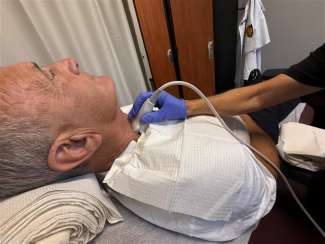 A man is lying down and a gloved hand holds an ultrasound device to his neck