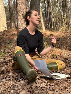 A female adult sits on the ground and is talking to a group.