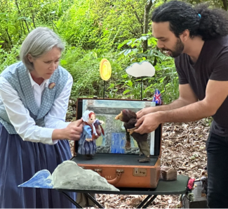 Two actors with tiny puppets performing in a suitcase set in the woods. 