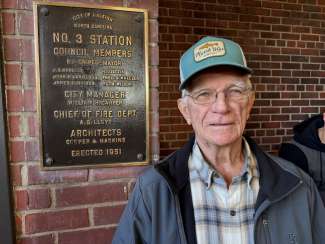 Retired Captain Wilson stands by bronze plaque bolted to brick wall at fire station 3