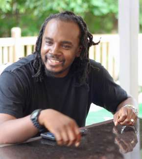Picture of a man with locks smiling and leaning in toward the camera.