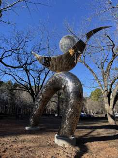 Large abstract metal sculpture with a curved, arch-like base and sweeping wing-shaped form supporting a circular disk, set outdoors among leafless trees under a clear blue sky.