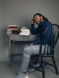 A young black man sitting thoughtfully at a grey book-covered desk, looking sideways at the camera.